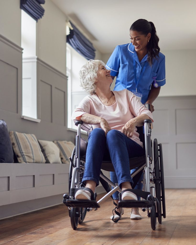 Senior Woman At Home Being Pushed In Wheelchair By Female Care Worker In Uniform